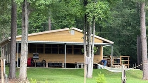 A lodge with a covered deck, a pond in the foreground, and trees surrounding the area.