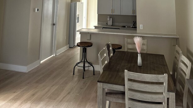 A spacious dining area with a wooden table and chairs, a kitchen in the background, and a ceiling with a grid pattern.