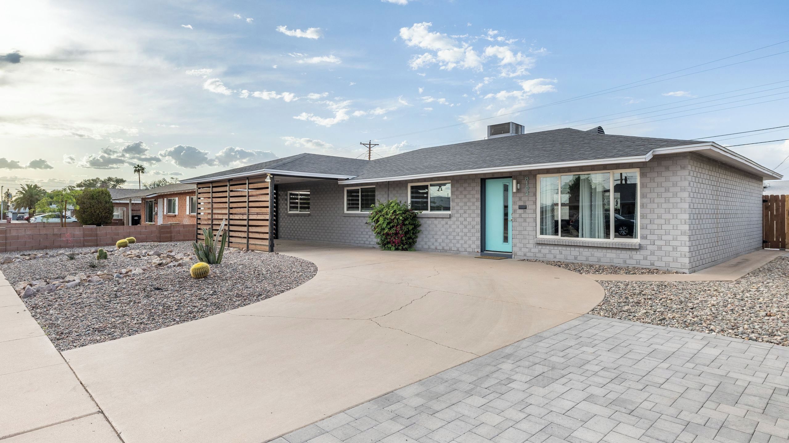 A residential house with a grey brick exterior, a blue door, and a small garden with cacti and shrubs.