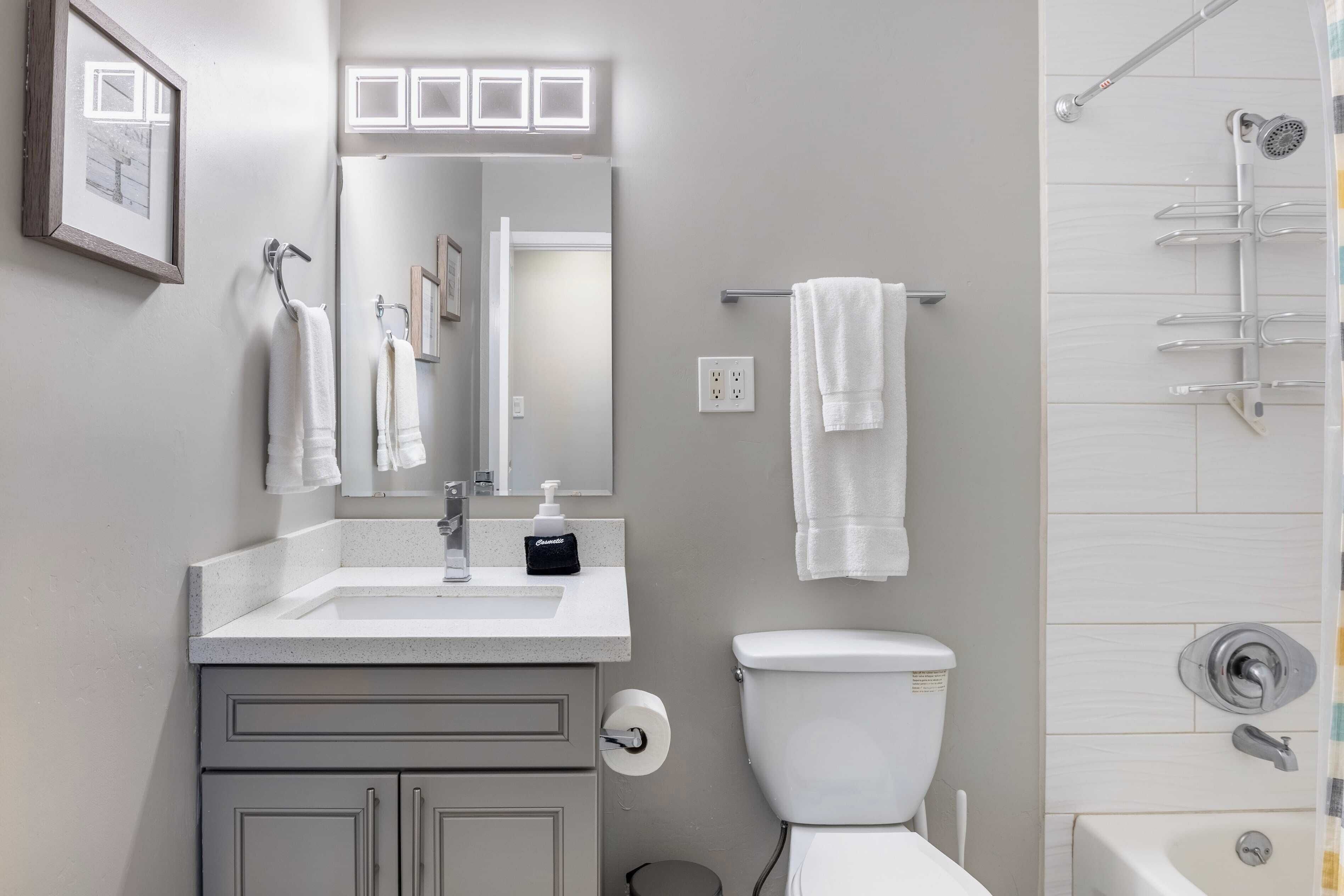 A modern bathroom with a white sink, mirror, and toilet. There are multiple white towels hanging on hooks.
