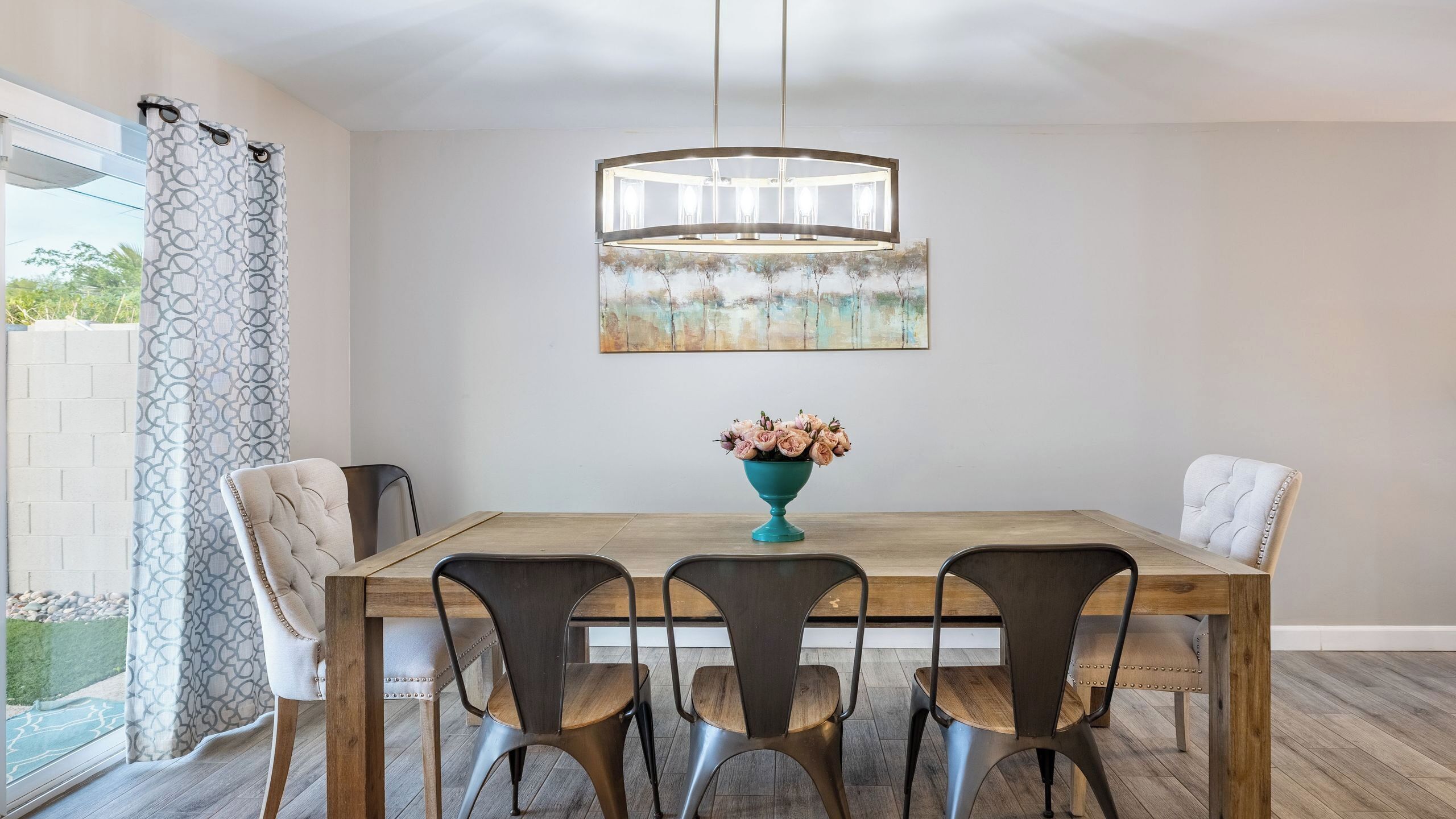 Zona pranzo con tavolo in legno, quattro sedie, un vaso color petrolio con fiori, un lampadario moderno e un grande quadro astratto appeso al muro.
