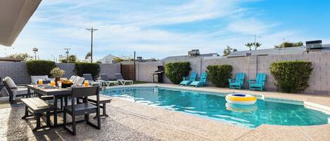 Un patio trasero con piscina, sillas de descanso, una mesa de comedor y una parrilla.