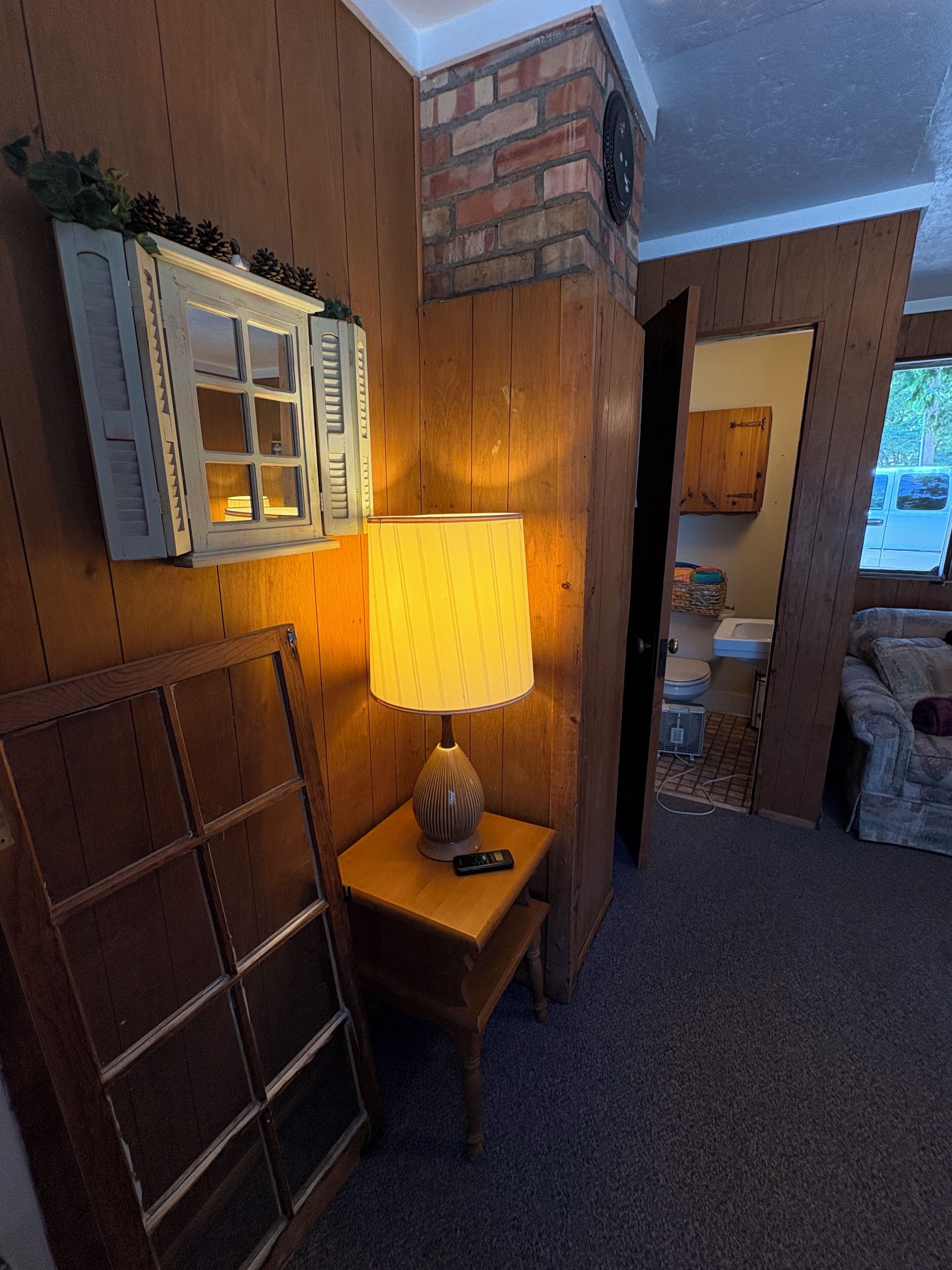 A wooden room with a lamp on a small table, a window with shutters, and a brick wall.