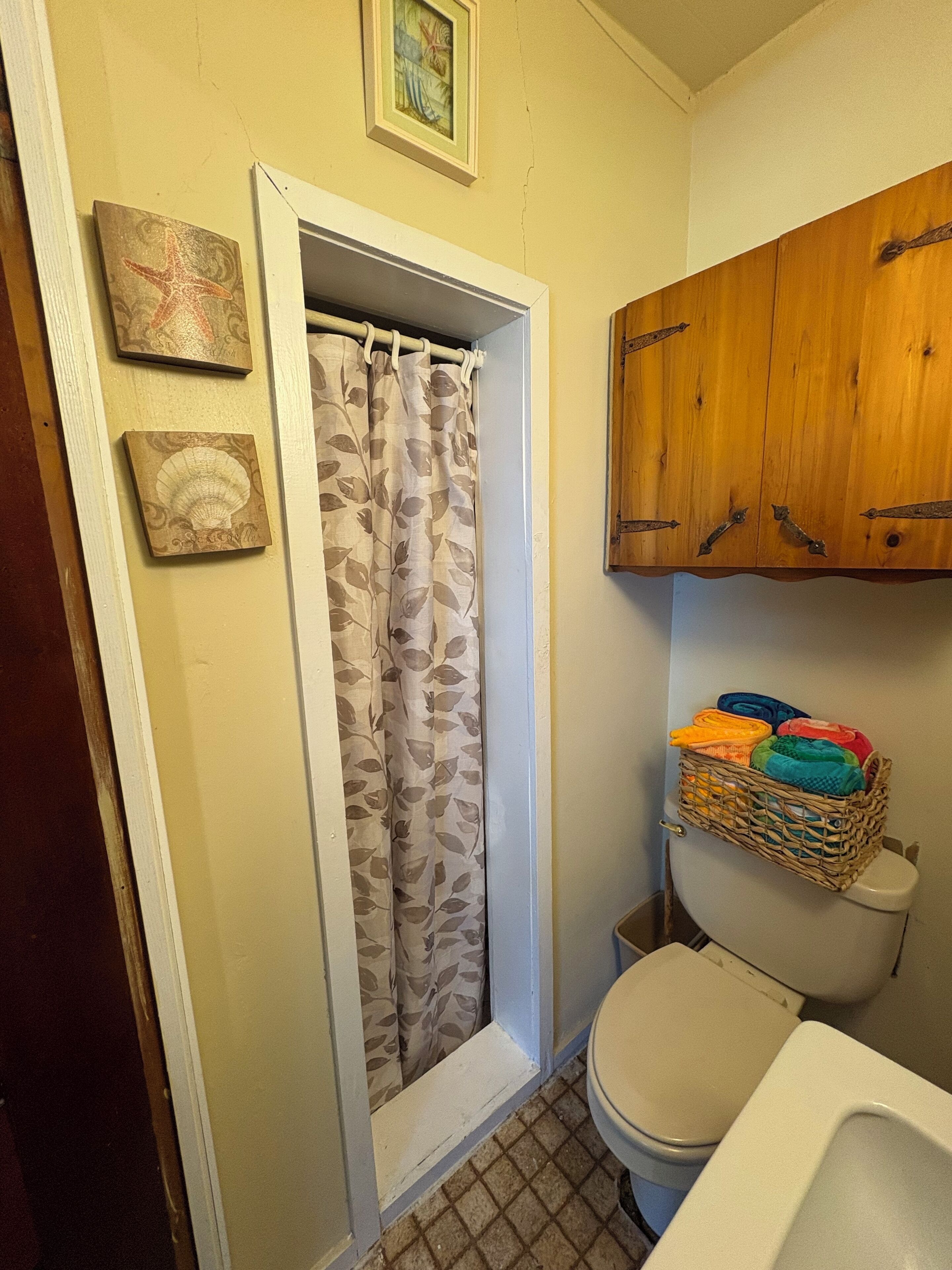 Une salle de bain avec des toilettes, un rideau de douche, des meubles en bois et un panier en osier contenant des serviettes.