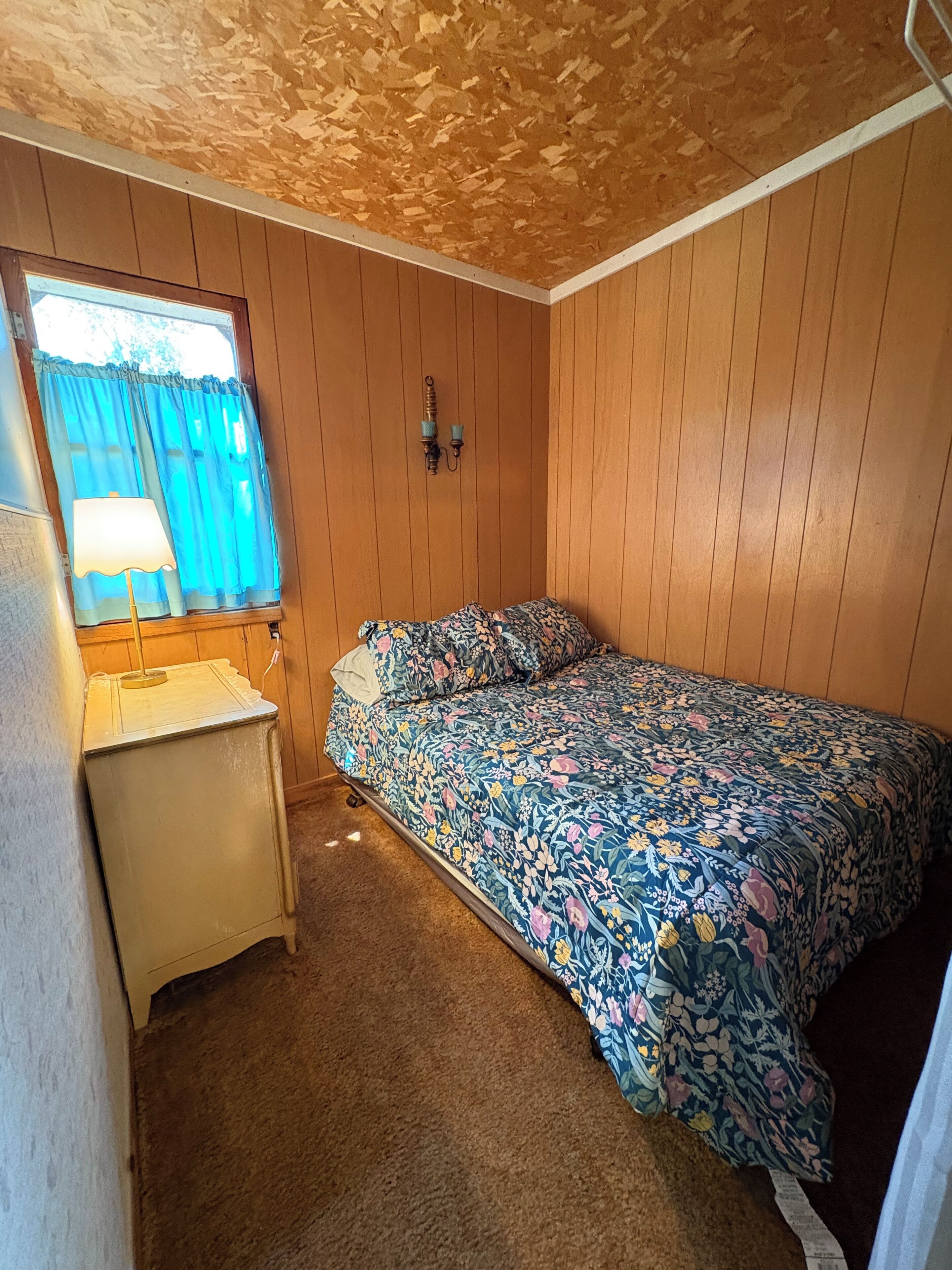 Et lille, hyggeligt rum med trævægge, en enkeltseng med blomstret sengetæppe, et lille bord med en lampe og et vindue med blå gardiner.