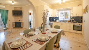 A dining area with a table set for four, a fireplace, and a kitchen in the background. - Corfu Hillside Villa – Terrace, Nature Walks, Sea Views (Agios Martinos)