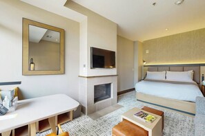 A hotel room with a bed, a TV mounted above a fireplace, a desk with a laptop, and a small table with a magazine.