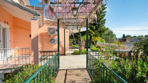 Une allée couverte, dotée d’un plancher en bois, mène à l’entrée d’un bâtiment, lequel est entouré de verdure et agrémenté d’une pergola couverte de plantes grimpantes en fleurs.