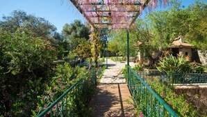 Un sentier de jardin bordé d’une pergola recouverte de fleurs roses, entouré d’une végétation luxuriante et d’un petit bâtiment doté d’un toit en tuiles.