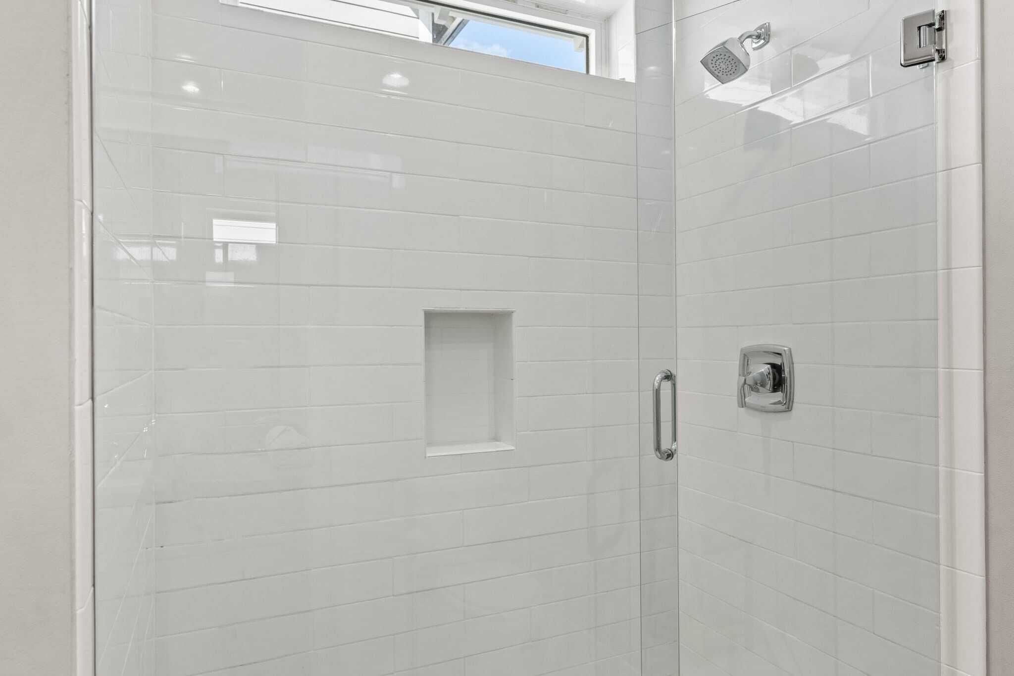 A modern bathroom with white tiled walls, a glass shower enclosure, and a small window above the shower.