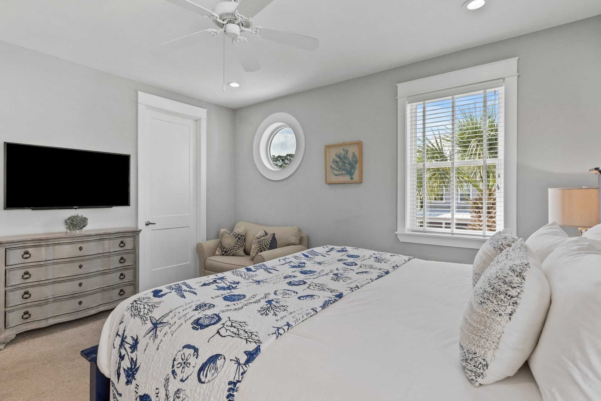 A bedroom with a bed, a dresser, a television, a ceiling fan, a window with blinds, and a framed picture on the wall.
