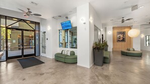 A modern lobby with a glass door entrance, ceiling fans, a flat-screen TV, and green seating areas. - Little Italy Inn - Limited Service Apart Hotel (San Diego)