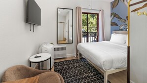 A bedroom with a bed, a ceiling fan, a TV, a mirror, a heater, a small table with a remote, and a window with curtains. - Little Italy Inn - Limited Service Apart Hotel (San Diego)