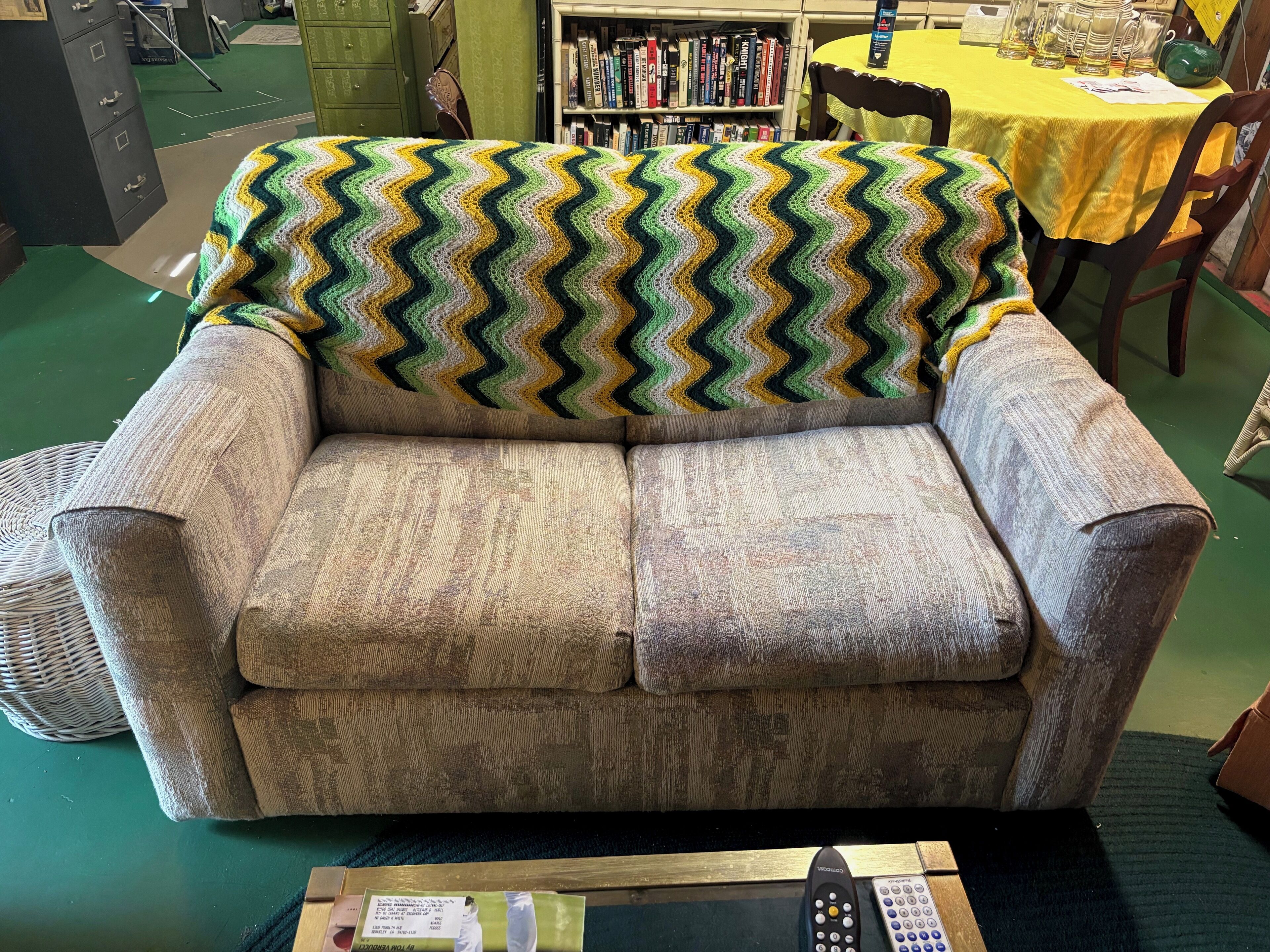 A grey sofa with a zigzag patterned blanket, a remote control, and a book on a table.