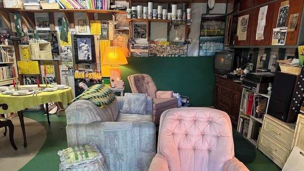 A room with a sofa, armchair, and a small table with a lamp. There are shelves with various items and a green wall in the background. - Charming baseball themed 2-story building with cable TV, wi-fi & stereo system. (Berkeley)