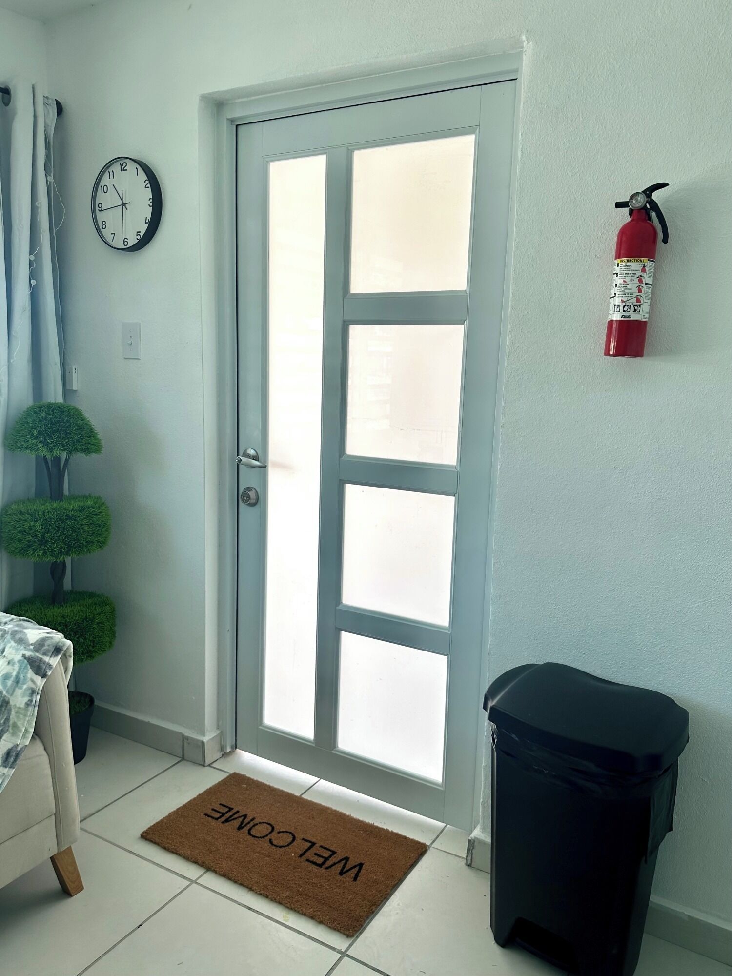 A room with a white door, a wall clock, a fire extinguisher, a welcome mat, and a trash bin.