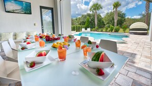 Un espace repas aménagé au bord de la piscine, avec une table dressée pour un repas, entouré d’une piscine et de palmiers.