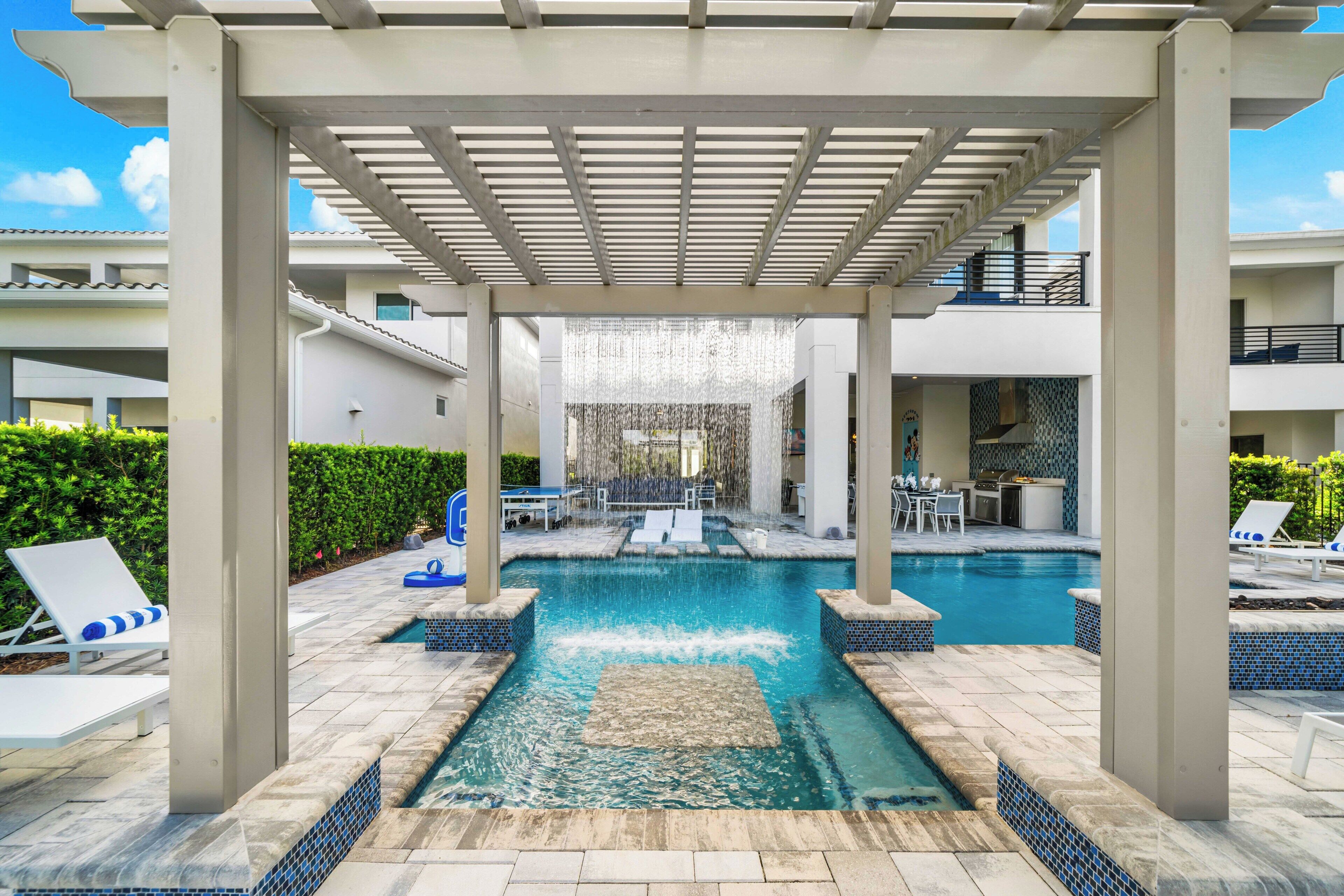 Ein Poolbereich mit einer Pergola darüber, Liegestühlen und einer kleinen Küchenzeile.