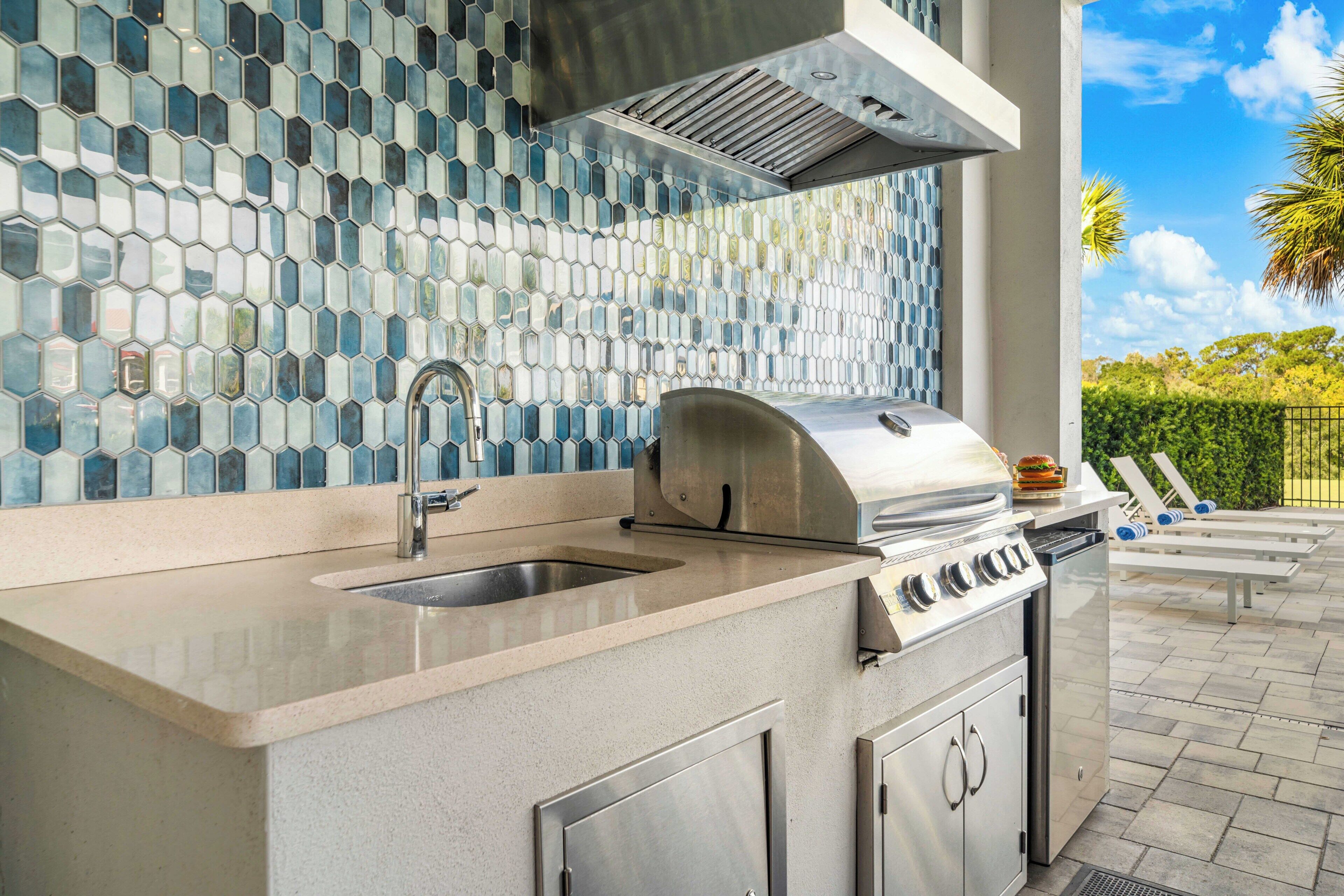 An outdoor kitchen with a stainless steel grill, a sink, and a backsplash with hexagonal tiles.