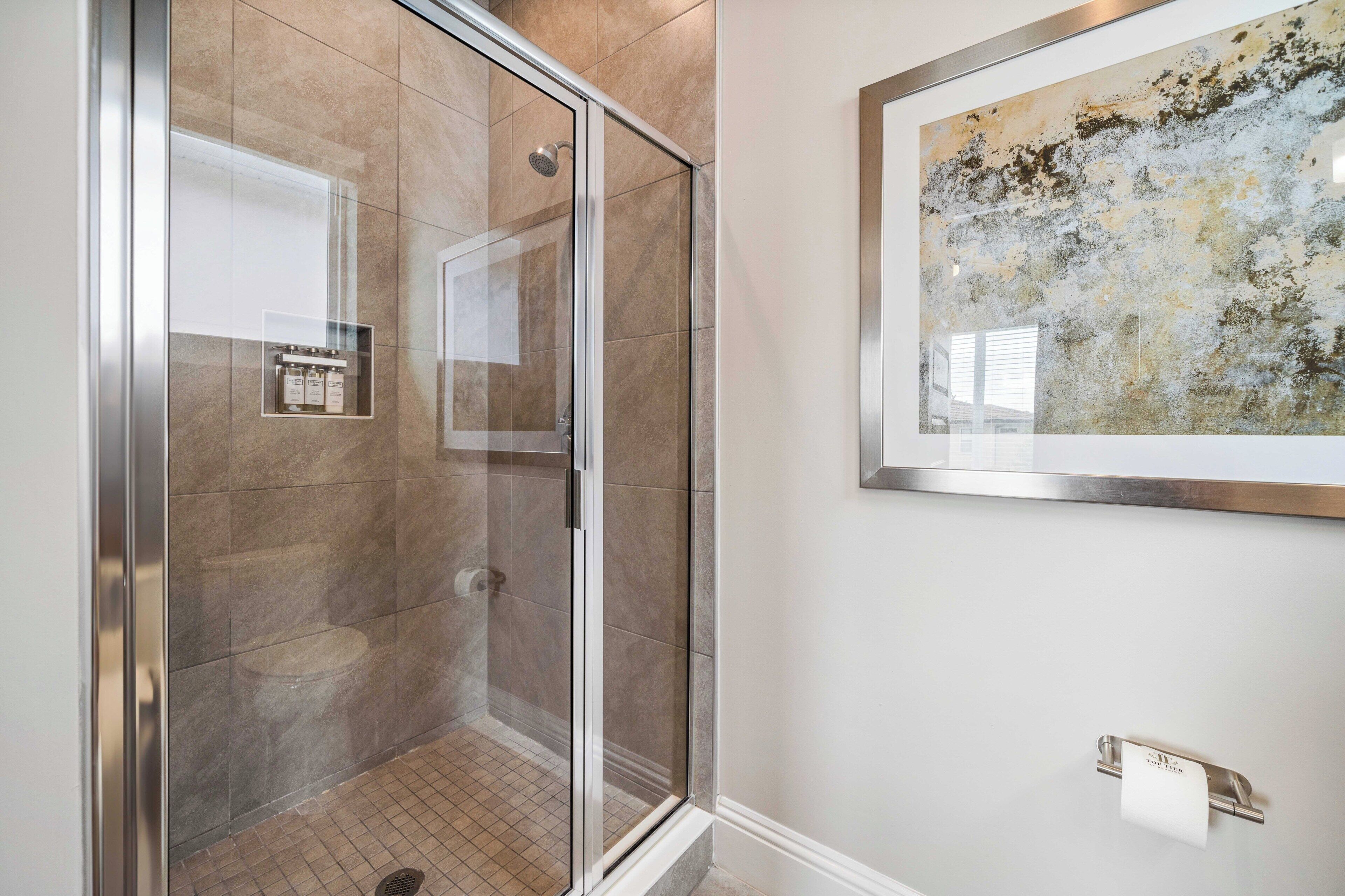 A glass-enclosed shower with a tiled floor, a framed abstract painting on the wall, and a towel dispenser.