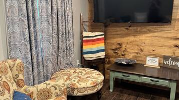Una sala de estar con un sofá, un televisor montado en una pared de madera, una mesita con un cuenco y un letrero, y cortinas con estampado.