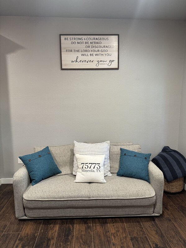Un sofá gris con dos almohadas azules y una almohada blanca con texto, frente a una pared lisa con una cita motivacional.