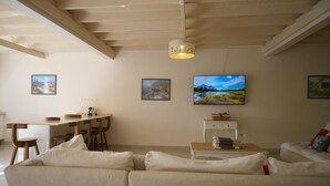 Ein Wohnzimmer mit einer Couch, einem Essbereich mit Stühlen, einem an der Wand befestigten Fernseher und gerahmten Bildern.
