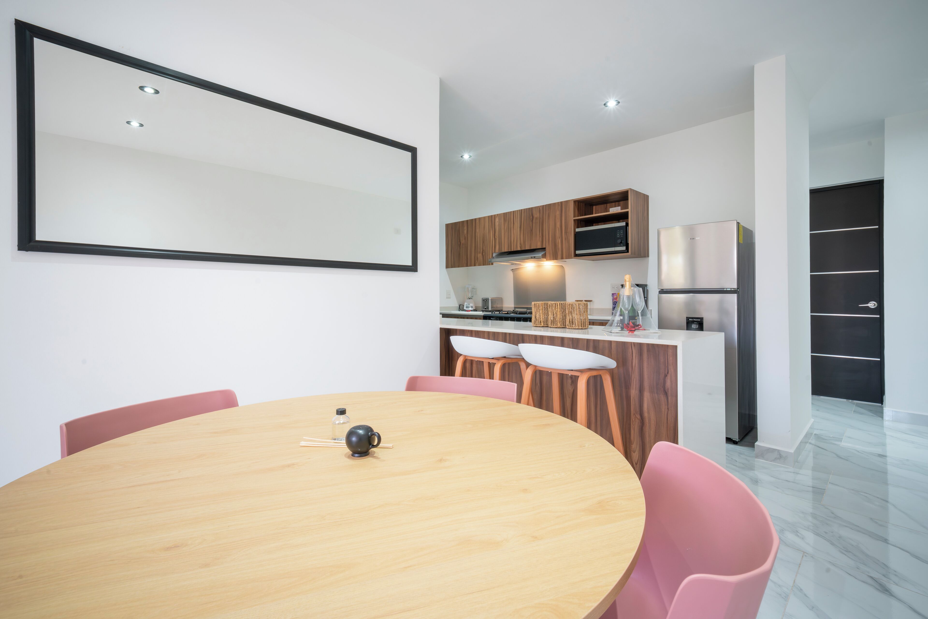 A modern kitchen with wooden cabinets, a stainless steel refrigerator, and a dining area with a round table and pink chairs.
