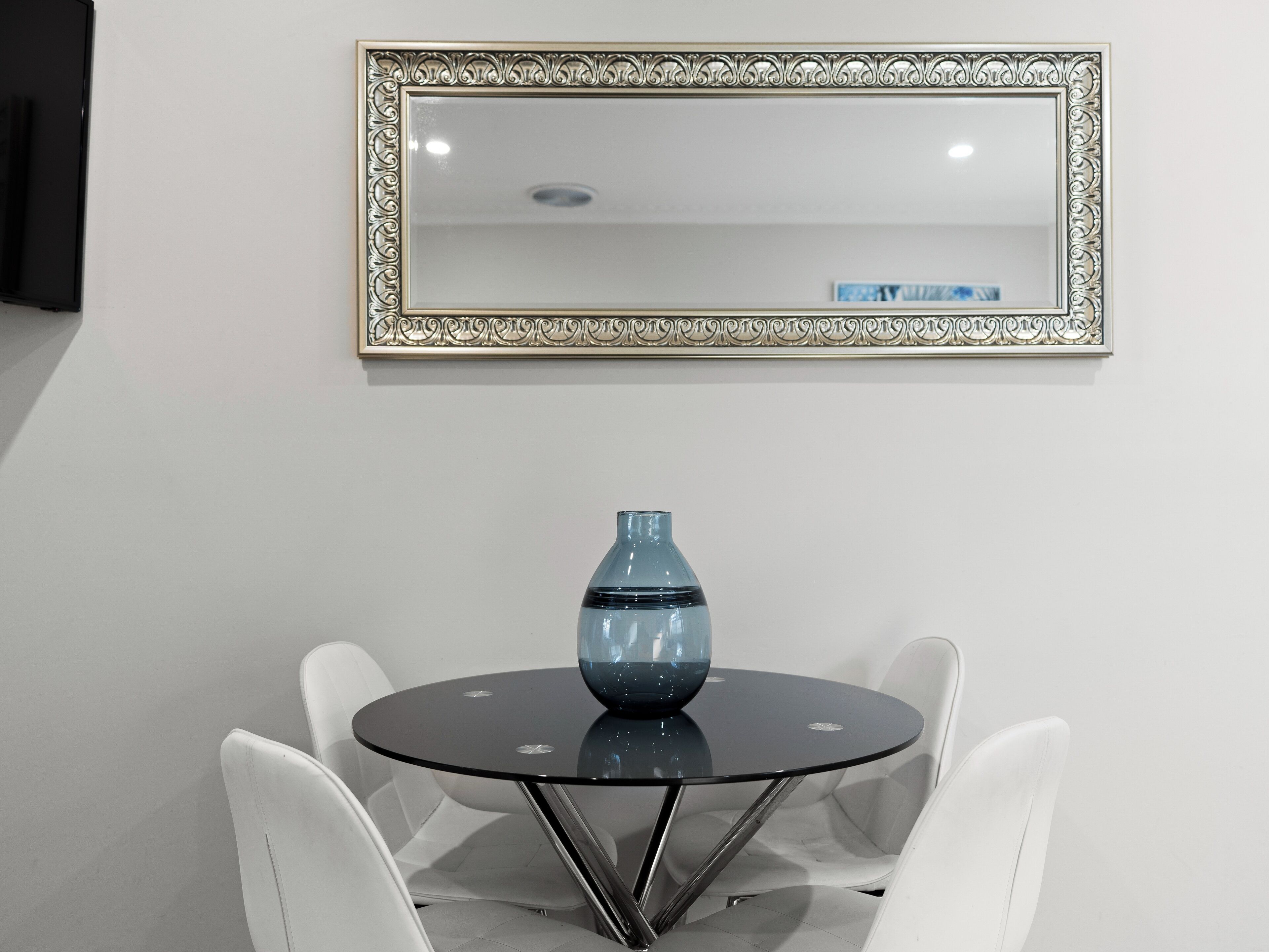 Uma mesa de vidro redonda com um vaso azul, rodeada por cadeiras brancas, sob um espelho decorativo.