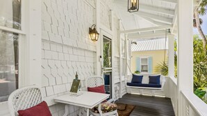 Ein überdachter Balkon mit weißen Holzwänden, einem weißen Tisch mit rotem Kissen und einer Schaukel mit blauen Kissen.