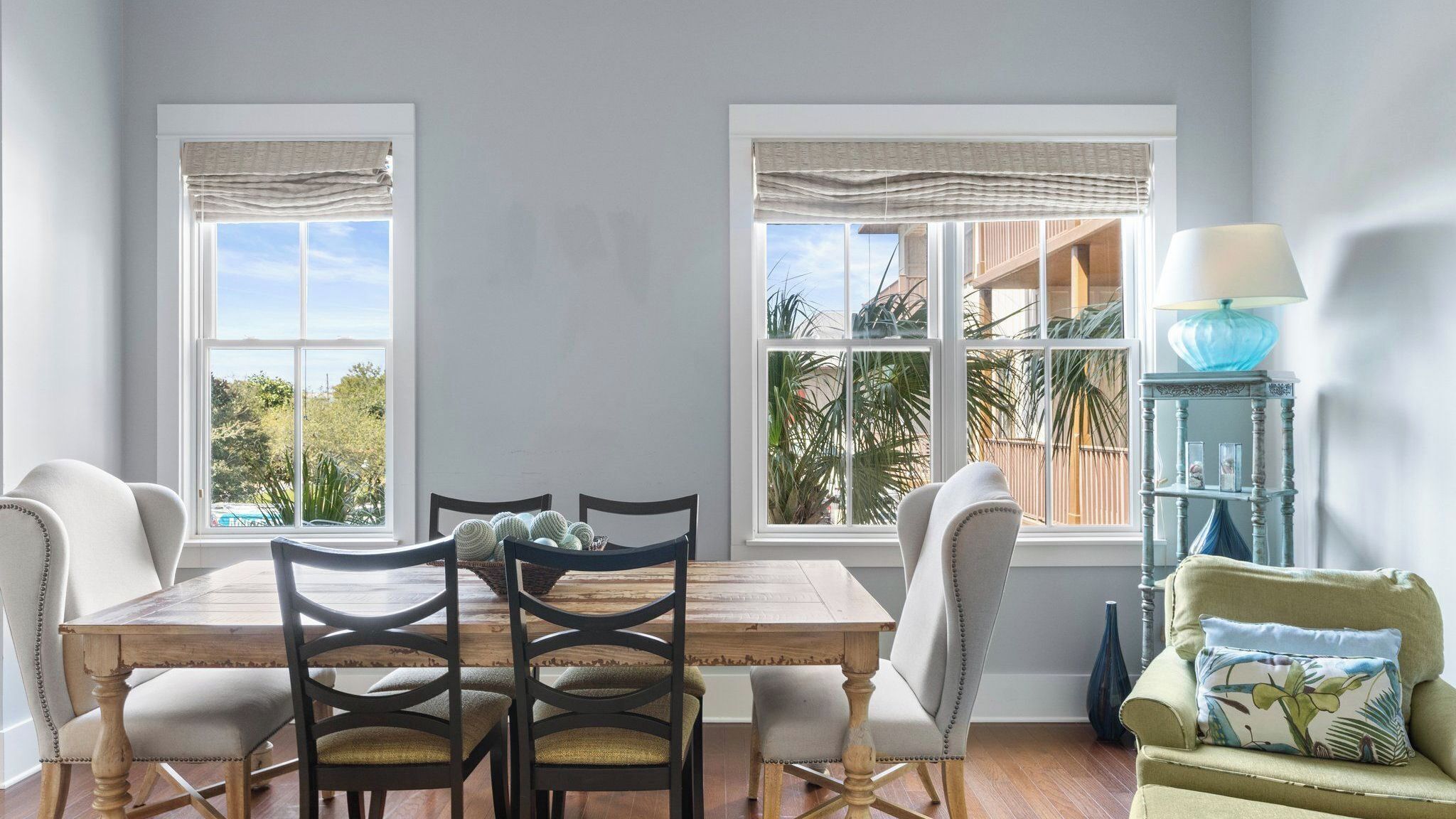 A dining area with a wooden table and chairs, two windows with blinds, a blue lamp, and a small blue vase.