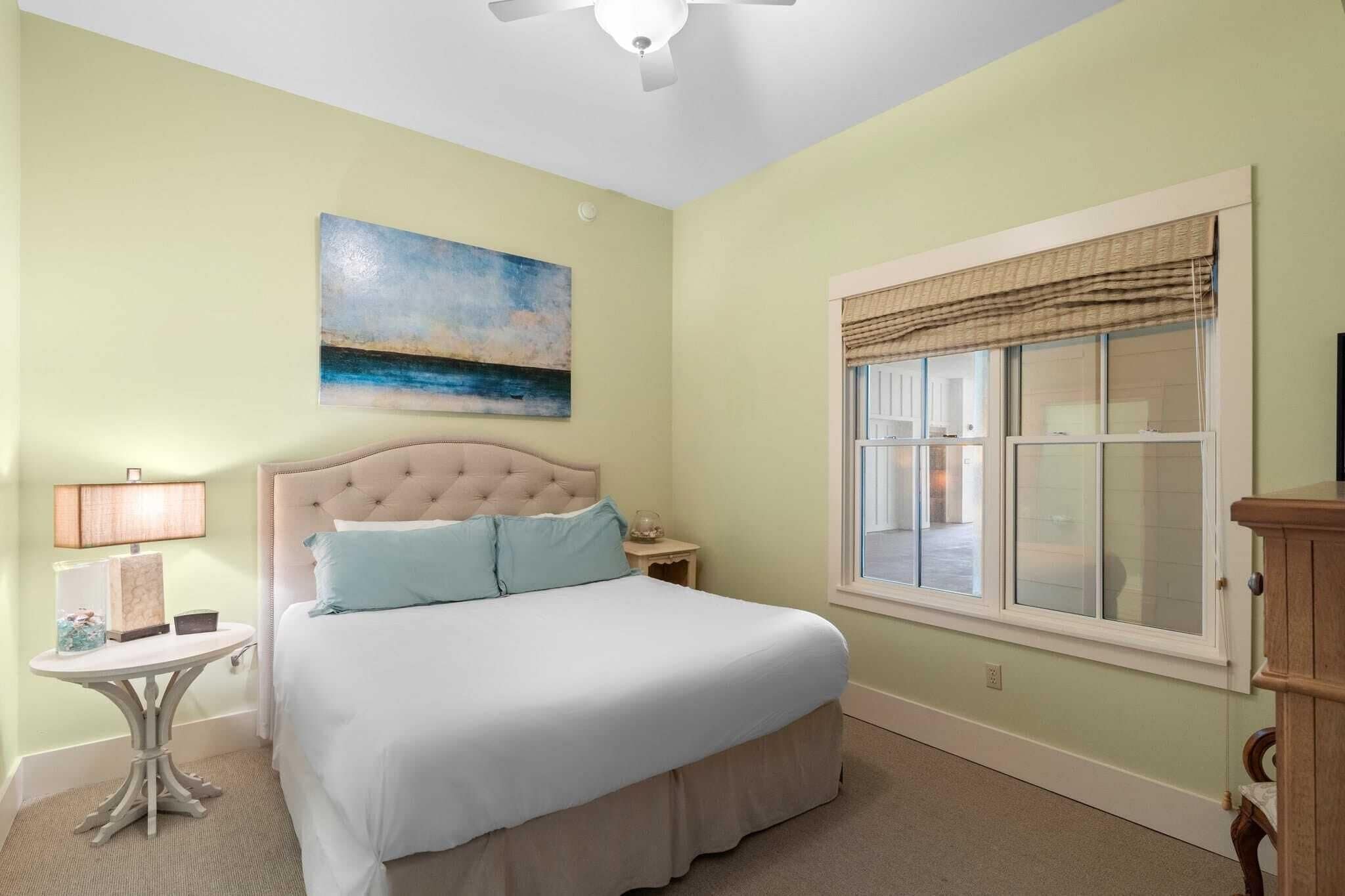 A bedroom with a bed, a nightstand, a lamp, a painting, and a window with blinds.