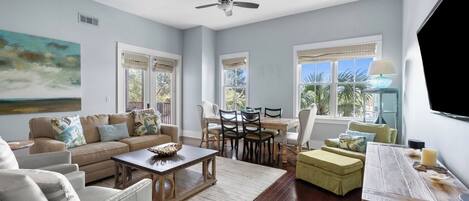 A spacious living room with a ceiling fan, a dining area with a table and chairs, and large windows with views of palm trees.