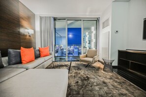 A modern living room with a sofa, armchair, and a glass table. A large window offers a city view at night.