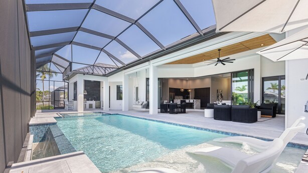 A modern indoor swimming pool with clear blue water, surrounded by white lounge chairs and a sunbed, under a large glass ceiling.