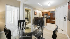A modern kitchen with stainless steel appliances, a dining area with a glass table, and a separate bathroom visible through an open door.