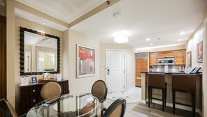 A dining area with a glass table and chairs, a kitchen with wooden cabinets and a microwave, and a door leading to another room. - SVS: Remodeled 1BD/2BA | Mountain View Balcony | Full Kitchen | MGM (Las Vegas)