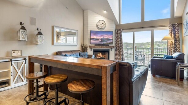 A modern living area with a bar, seating, a fireplace, and a large window offering a view. - Family-Friendly, Fishing, & Fun w/ 2 Pools, Views, Boat Slip & Launch (Camdenton)