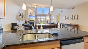 A modern kitchen with granite countertops, a double sink, and a dishwasher. There is a dining area with a table and chairs, and a view of a lake through large windows. - Family-Friendly, Fishing, & Fun w/ 2 Pools, Views, Boat Slip & Launch (Camdenton)