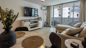 Ein modernes Wohnzimmer mit Deckenventilator, Flachbildfernseher, einer Couch, einem Esstisch mit Platzsets und einem großen Fenster mit Vorhängen.