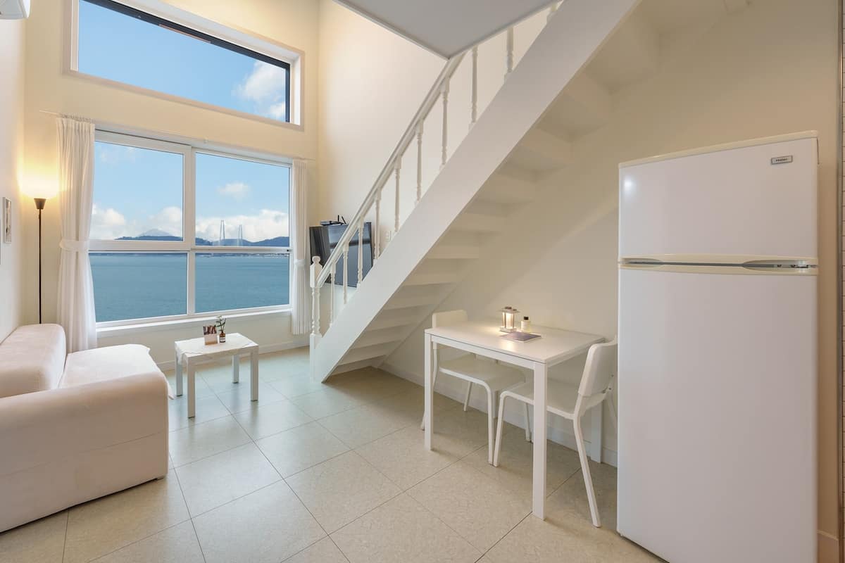 A modern, well-lit room with a large window offering a scenic view of the sea and mountains. It includes a sofa, a small table, and a refrigerator.
