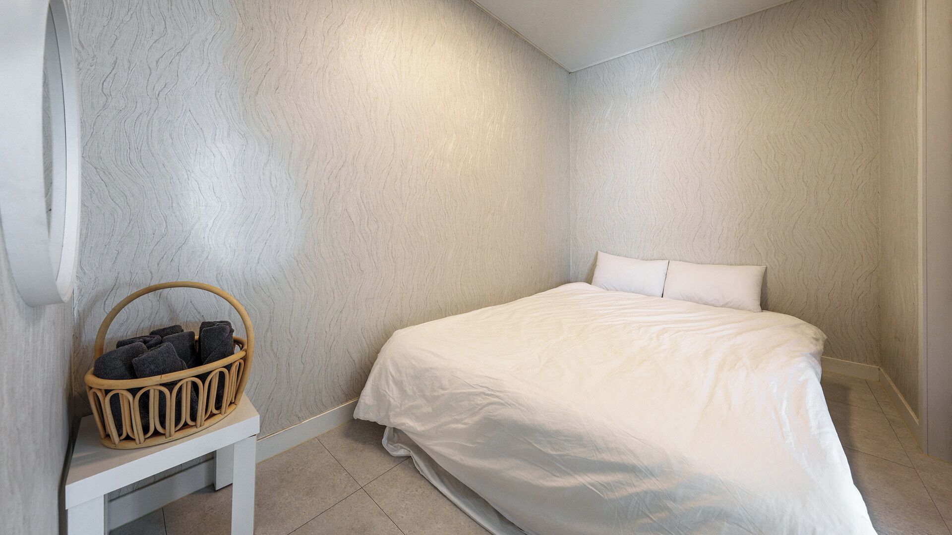 Une chambre à coucher avec un lit, un oreiller blanc, une table de chevet blanche et un panier en osier contenant des serviettes.