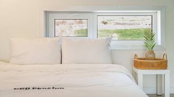 Un dormitorio con cama, almohadas, una mesita de noche con una planta y una ventana que da a una zona verde.