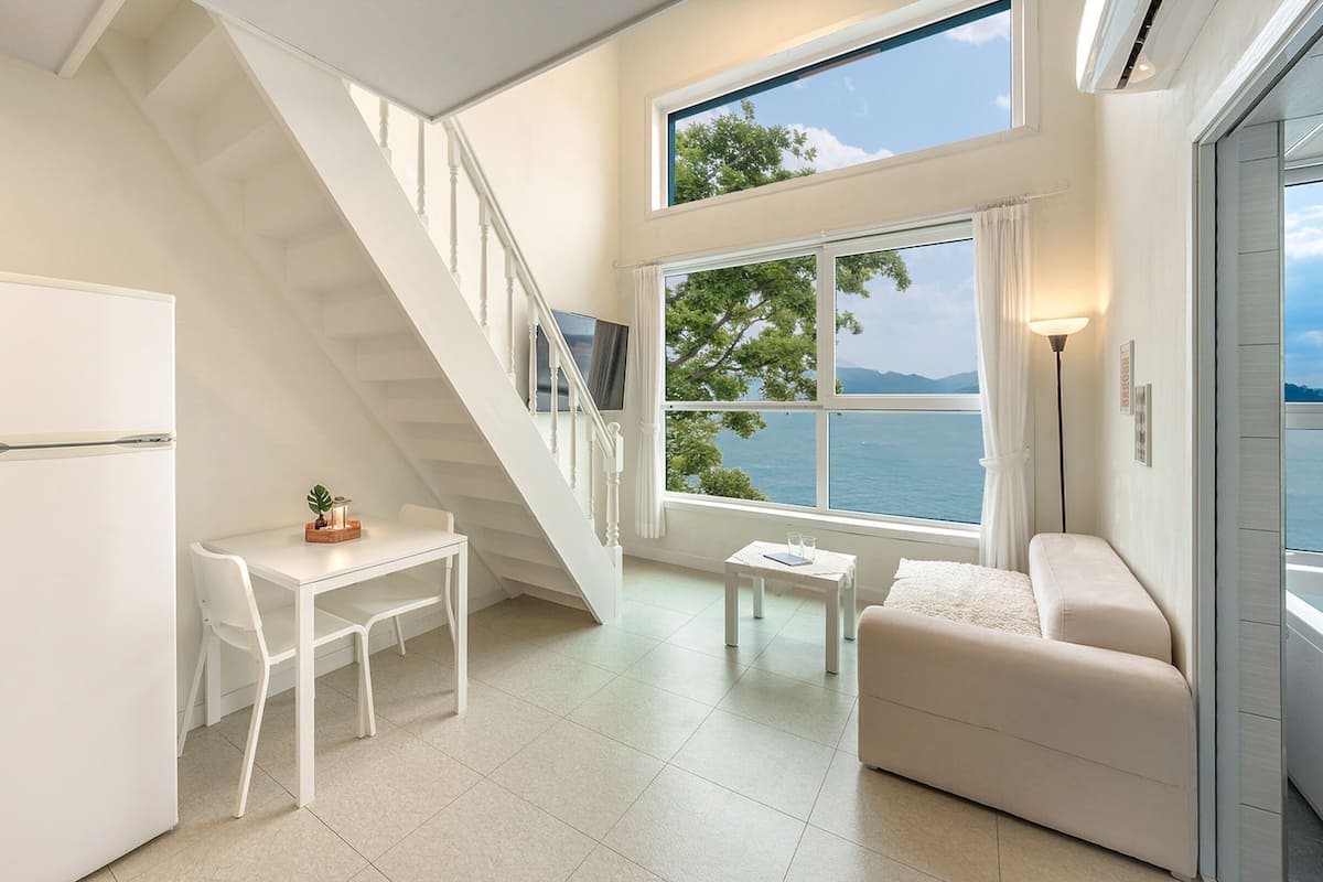 A compact living space with a kitchen, dining area, and seating by a large window overlooking a body of water.