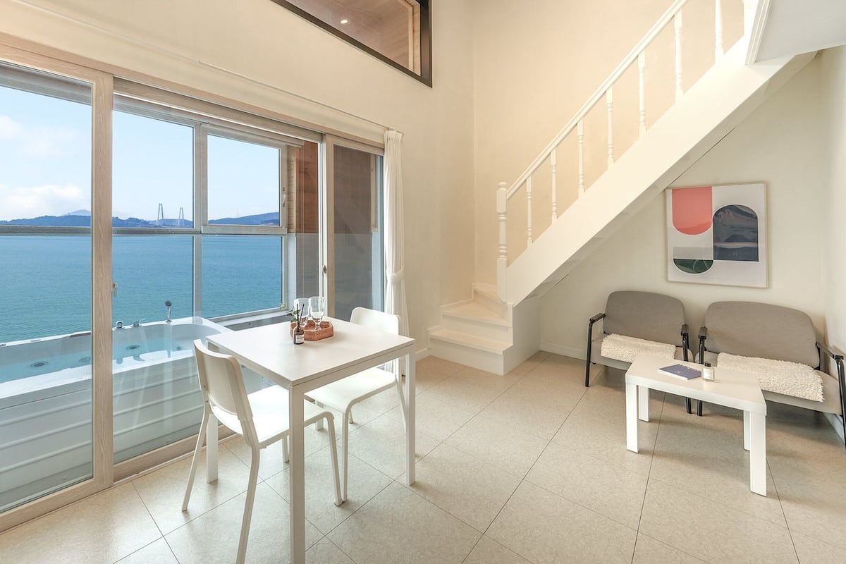 A modern room with a large window offering a sea view, a white table with chairs, a sofa, and a staircase.