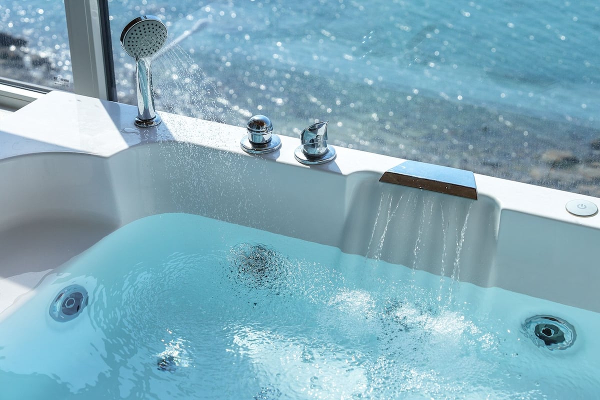 A bathtub with multiple faucets and a view of the sea through a window.