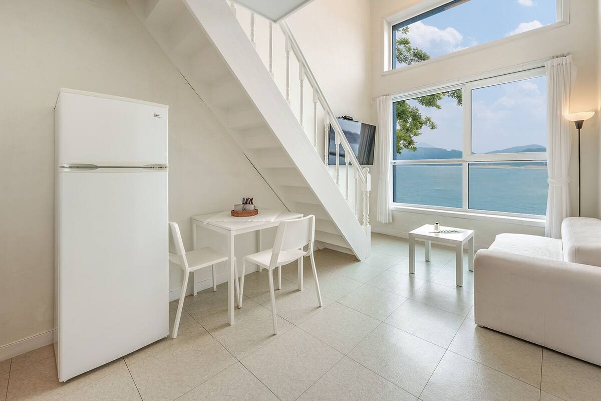 Un salón moderno y minimalista con un sofá blanco, una mesa de comedor con sillas, una mesita blanca y un refrigerador blanco. Una escalera conduce a un balcón con vista al mar.