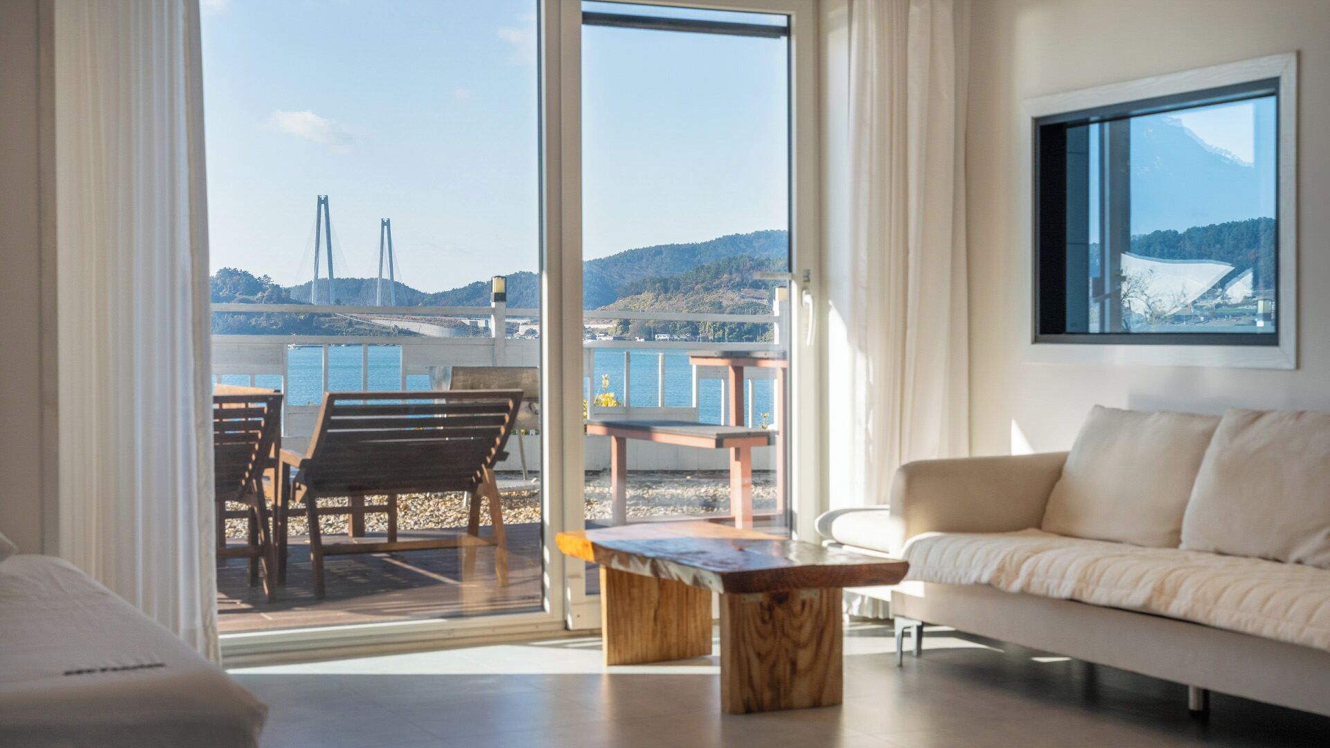 A modern living room with a sofa, a coffee table, and a view of a bridge and hills through large windows.