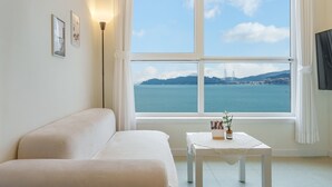 A room with a white sofa, a small table with a vase and a bottle, and a large window with a view of a body of water and a bridge. - Yeosu Jangsuri Poolvilla&spa Pension (Yeosu)