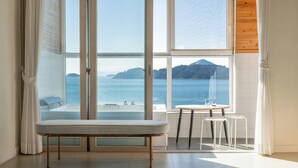 A room with a bench, two stools, a table, and a large window offering a view of the sea and a mountain. - Yeosu Jangsuri Poolvilla&spa Pension (Yeosu)
